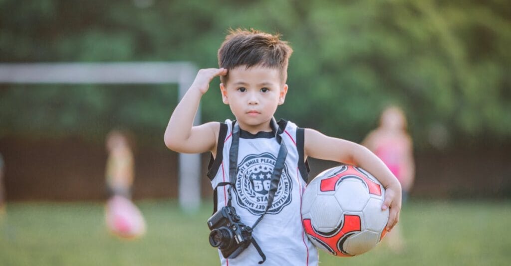 A young boy holding a soccer ball and camera stands confidently on a sunny day.