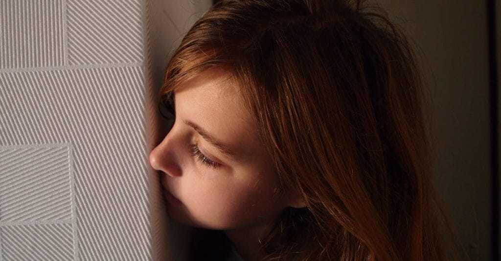 A young girl with blonde hair peeking playfully from behind a textured wall indoors.