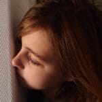 A young girl with blonde hair peeking playfully from behind a textured wall indoors.