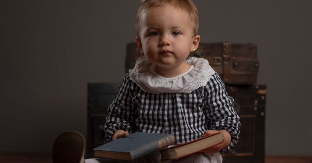 Charming baby in checkered dress sitting indoors with books and luggage trunk.