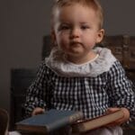 Charming baby in checkered dress sitting indoors with books and luggage trunk.