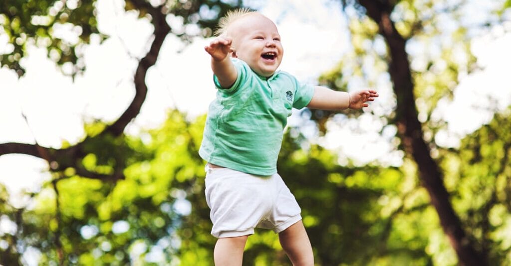 Happy baby wearing a green shirt being tossed in the air on a sunny day in the park.