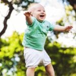 Happy baby wearing a green shirt being tossed in the air on a sunny day in the park.