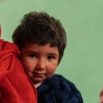 Two Afghan children in traditional clothing, captured in a candid moment indoors.