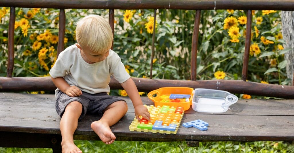 Toddler enjoys a puzzle toy on a wooden bench surrounded by summer flowers.