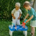 Two young boys engaged with a play table in a sunny garden setting, enjoying a fun summer day.