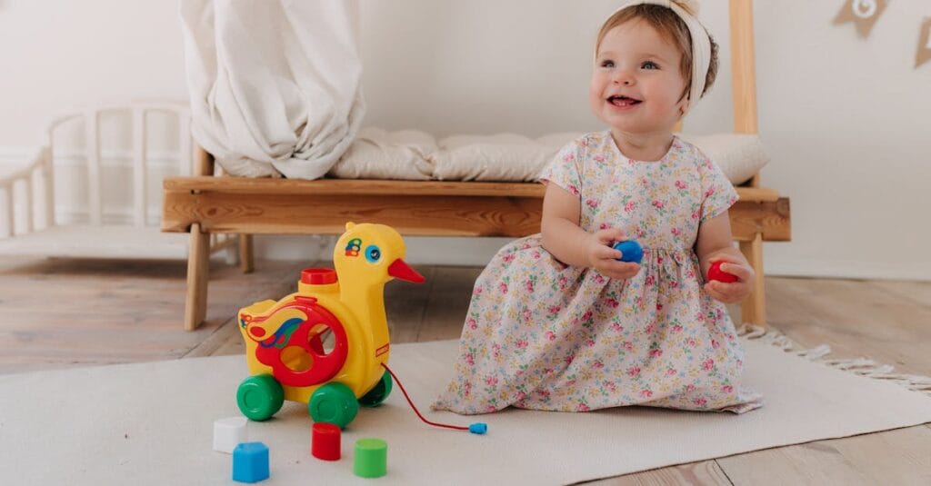 A smiling baby girl plays with a colorful toy in a cozy nursery setting.