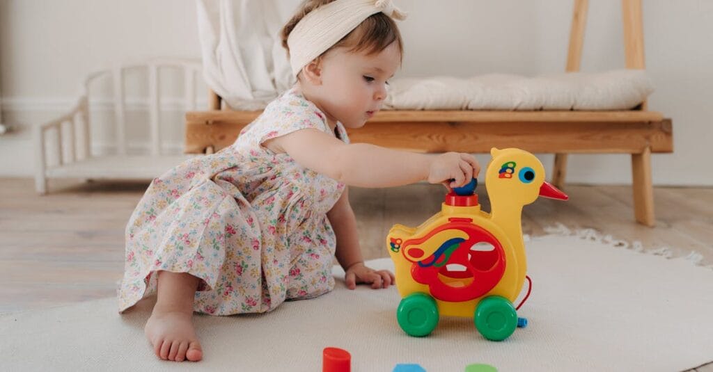 Cute baby girl in a floral dress playing with a colorful duck toy indoors.