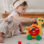 Cute baby girl in a floral dress playing with a colorful duck toy indoors.