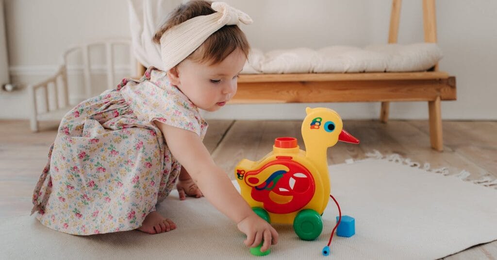 Cute baby girl playing with a colorful duck toy on a white rug in a cozy room.