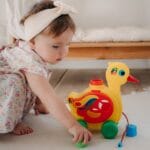 Cute baby girl playing with a colorful duck toy on a white rug in a cozy room.