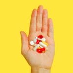 Close-up of an adult hand holding different types of pills against a vivid yellow background.