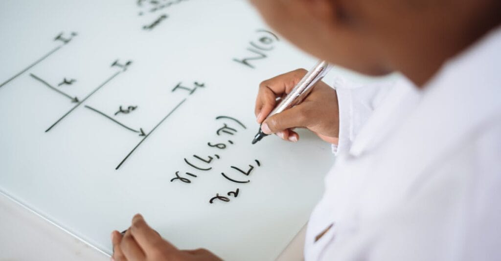 From above anonymous ethnic student wearing uniform and solving problem in chemistry while writing formula on white table in lab