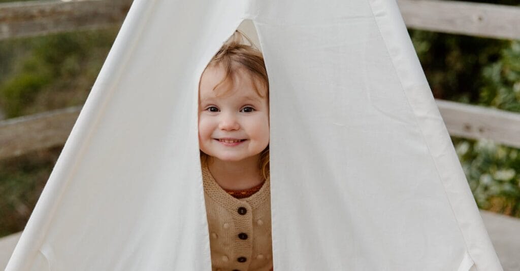 Smiling child peeks out from a cozy tent, exuding joy and playfulness in a natural setting.