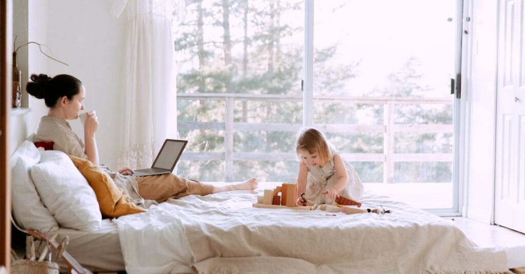 Concentrated female drinking coffee while working on project on laptop and daughter sitting on bed playing with constructor in bedroom
