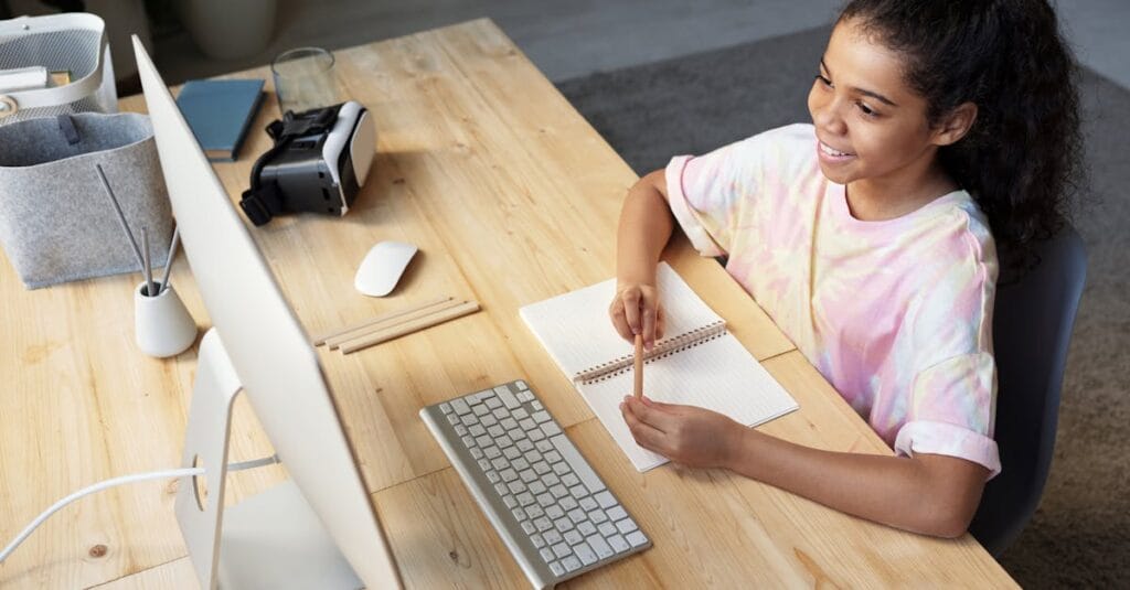 A young girl smiling while studying online at home, using a computer and taking notes.