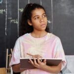 A teenager in a pink shirt ponders while holding a notebook in a classroom setting.