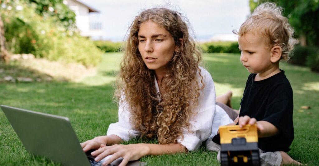 Woman working on a laptop while lying on grass with her child nearby playing.