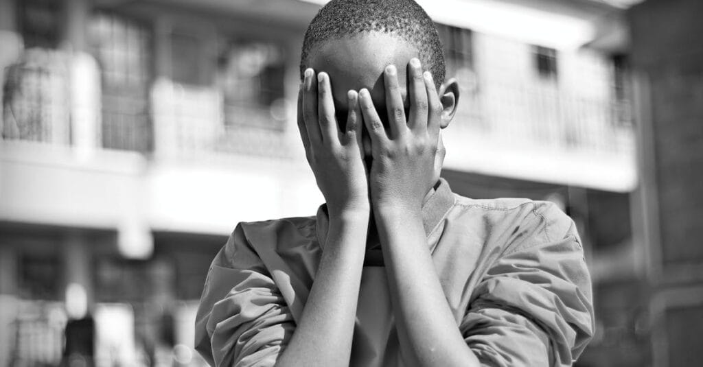 Black and white image of a child covering their face with hands outdoors in Nairobi.