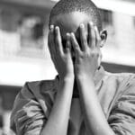 Black and white image of a child covering their face with hands outdoors in Nairobi.