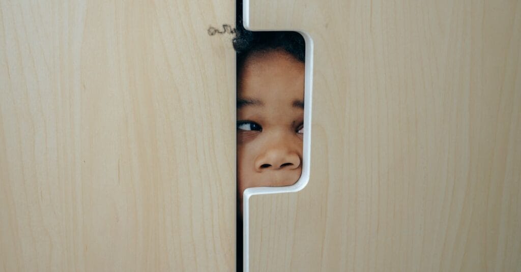 Anonymous funny little ethnic child hiding in wardrobe and peeping through hole while playing at home