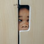 Anonymous funny little ethnic child hiding in wardrobe and peeping through hole while playing at home