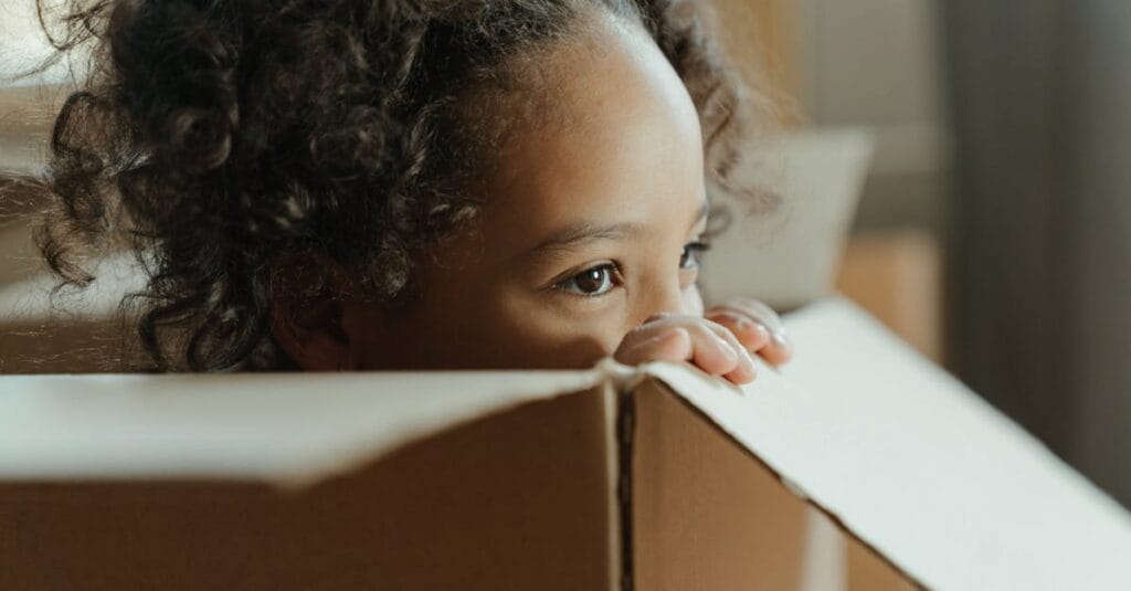 A charming portrait of a child with curly hair peeking out from a cardboard box indoors.