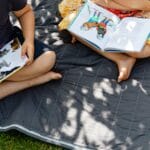 Two kids enjoy reading illustrated books on a picnic blanket in a sunny outdoor setting.