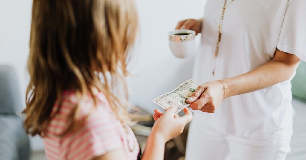 A woman hands a dollar bill to her daughter while holding a coffee cup, indoors.