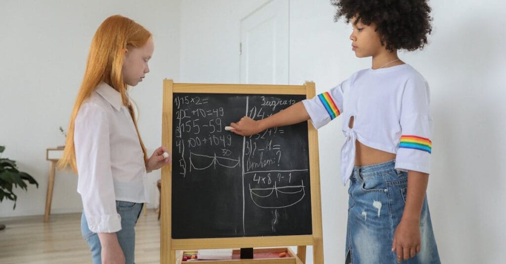 Two girls solving math equations on a chalkboard in a bright classroom setting.