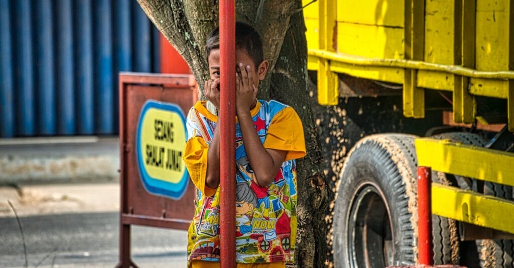 A child playing hide and seek beside a tree and a vibrant yellow truck in Purwakarta, Indonesia.