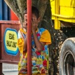 A child playing hide and seek beside a tree and a vibrant yellow truck in Purwakarta, Indonesia.