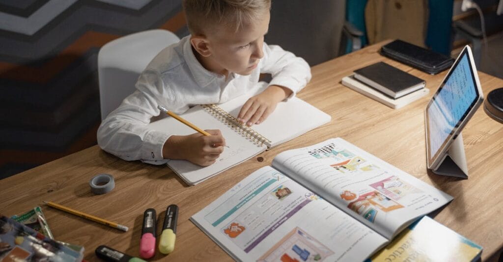 A focused young boy engages in distance learning with a tablet, books, and notebooks on a desk.