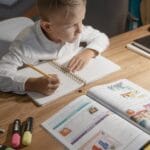 A focused young boy engages in distance learning with a tablet, books, and notebooks on a desk.
