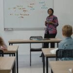 A diverse group of students in an English class with a smiling teacher at the whiteboard.