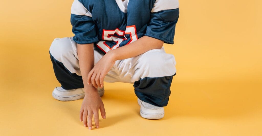 Young child in football gear squatting on a yellow background, showcasing sporty attire and enthusiasm.