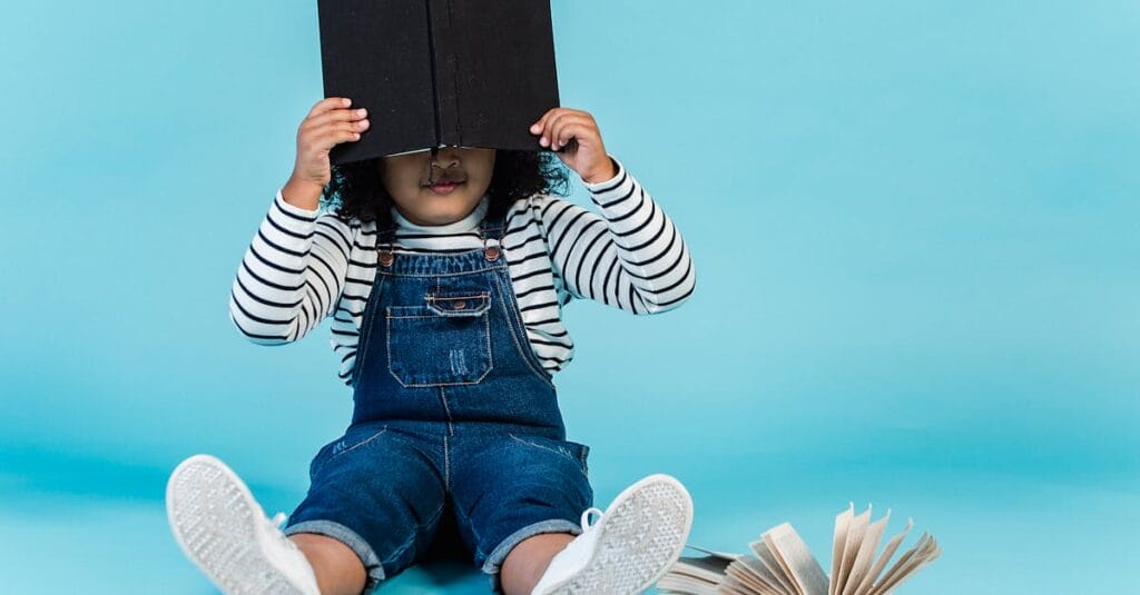 Unrecognizable little African American girl in trendy clothes sitting on floor with textbooks and covering face with book against blue background