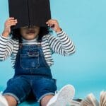 Unrecognizable little African American girl in trendy clothes sitting on floor with textbooks and covering face with book against blue background