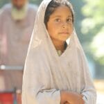 A young girl stands outdoors, looking at the camera with a serene expression in traditional attire.