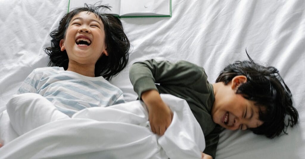 Two happy children laughing together on a cozy bed in the morning light.