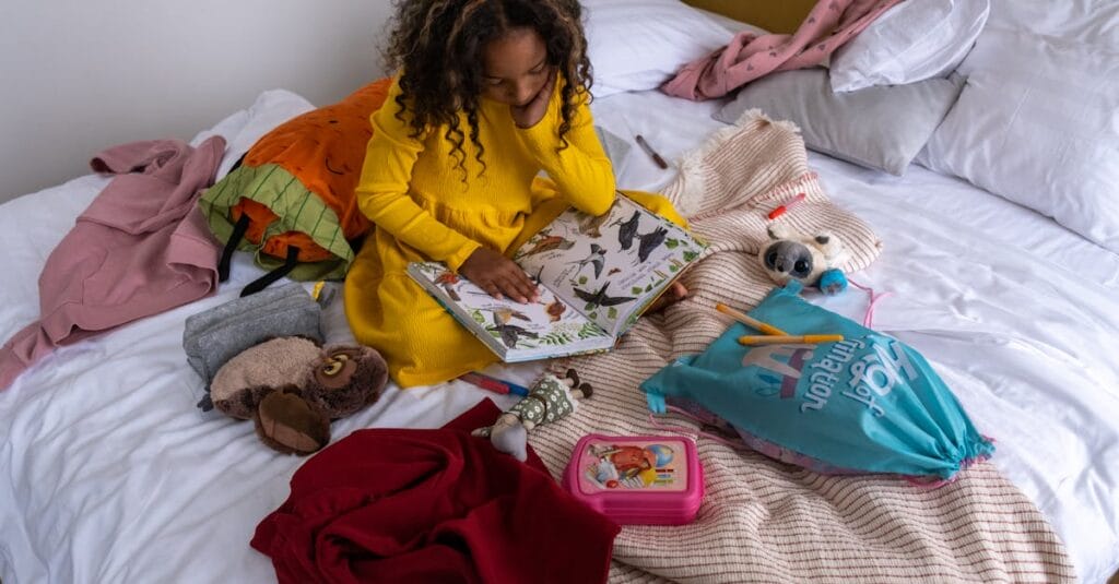 Young girl reading a book on a bed, surrounded by toys, clothes, and personal items. Cozy indoor setting.