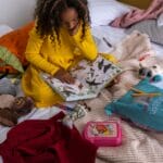 Young girl reading a book on a bed, surrounded by toys, clothes, and personal items. Cozy indoor setting.
