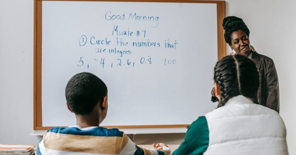 Cheerful African American female math teacher in formal clothes standing near whiteboard and explaining lecture task to multiracial pupils