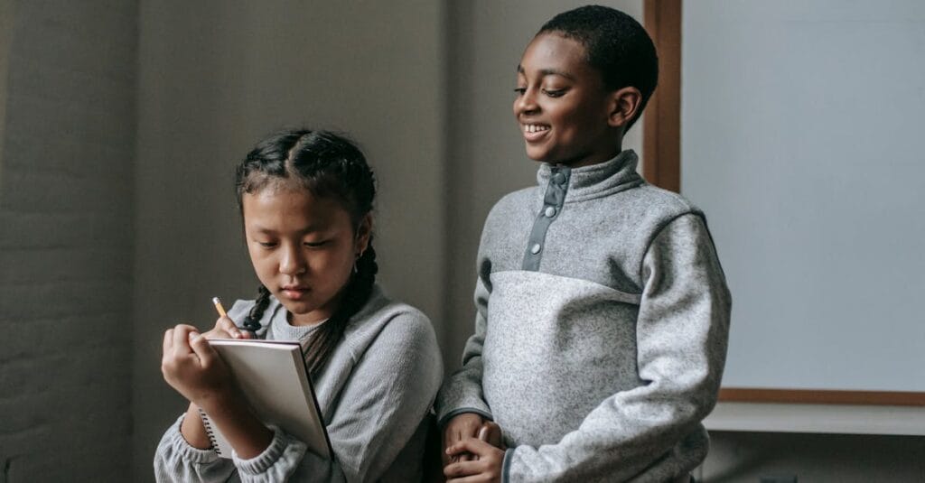 Two children engaged in a classroom activity, one smiling and the other taking notes.