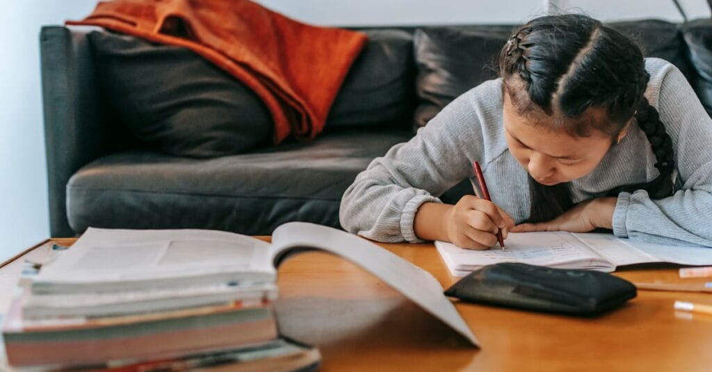A young Asian girl with pigtails diligently completing homework at home on the sofa.