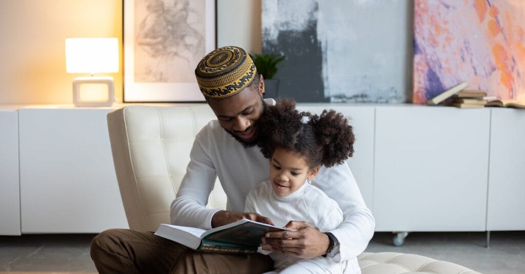 Positive young African American father in hat reading book to cute little daughter sitting on armchair at home