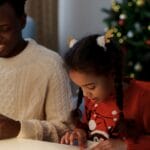 Father and daughter crafting together by the Christmas tree, enjoying holiday bonding time.