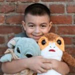 A cheerful child hugs Grogu and Porg plush toys against a rustic brick wall background.