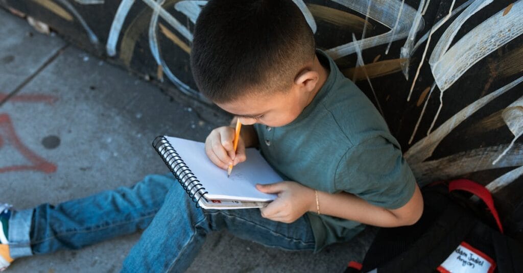 Child sitting on sidewalk, concentrating on writing in notebook with pencil, surrounded by urban graffiti.