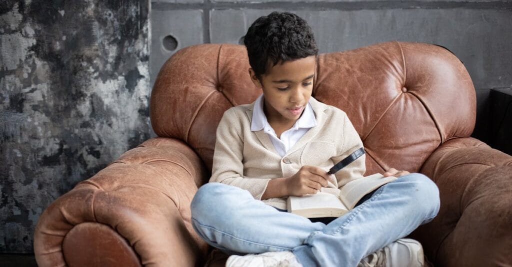 Full body of curious Arabian schoolboy sitting on comfortable armchair with crossed legs and reading book with magnifier at home